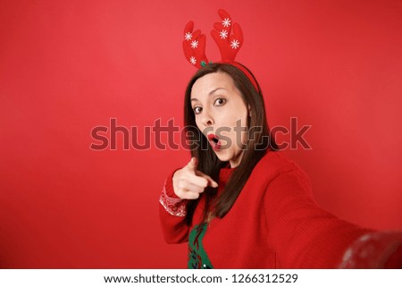 Close up selfie shot of amazed Santa girl in decorative deer horns pointing finger on camera, keeping mouth wide open isolated on red background. Happy New Year 2019 celebration holiday party concept