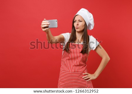Housewife female chef cook or baker in striped apron, white t-shirt, toque chefs hat isolated on red wall background. Smiling fun woman doing selfie shot on mobile phone. Mock up copy space concept