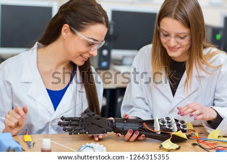 Two women scientists examining mechanical hand in laboratory. Female students, wearing in uniform, studying structure of prototype of mechanical plastic hand robot that can using to prosthetic people. Royalty-Free Stock Photo #1266315355
