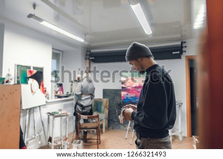 Portrait of a young artist standing in the studio and using the internet on a smartphone,against the background of paintings,easels and the painter paints a picture.Creative atmosphere in art studio
