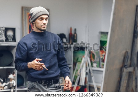 Portrait of a concentrated busy artist stands with a brush in his hands and looks at the picture. Painter in a cozy studio working on a new painting. Work as a hobby.