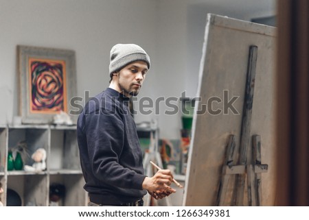 Busy man artist with a brush in his hand picks up an oil painting in a cozy studio. Talented young painter paints a painting on canvas. Painting Concept. Work as a hobby.
