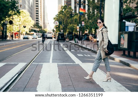 young office lady holding coffee confident walking on road wearing high heels and sunglasses. businesswoman on zebra crossing. rainbow flag hanging on street in background local people riding bike