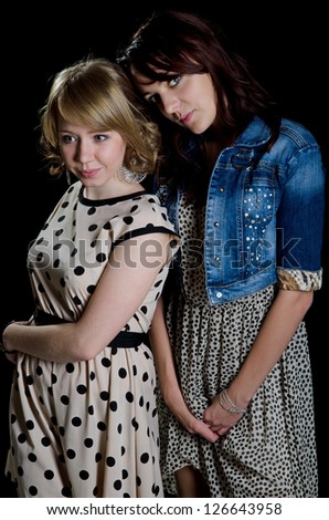 Two attractive trendy young woman in stylish dresses posing close together on a dark studio background