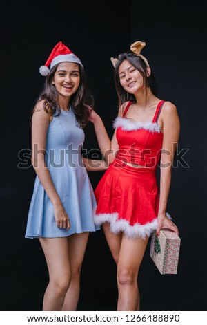 Two young Asian girls (one Indian, one Chinese) exchange Christmas gifts. One is wearing a red Santarina outfit and the other a blue dress with a Santa hat. They are both smiling happily. 