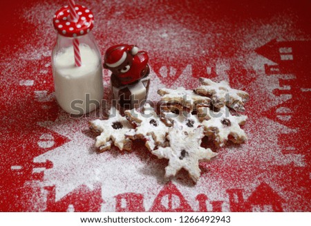 Snowflakes shaped Christmas cookies on a red background with powdered sugar silhouettes of houses under the cookies and a milk bottle with straw in it, and a chocolate bear shape dessert 1