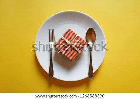 New Year celebrations theme red and brown striped gift box with white tied rope in a bow on white plate with fork and spoon over yellow background. Holidays well wishing concept. (selective focus)