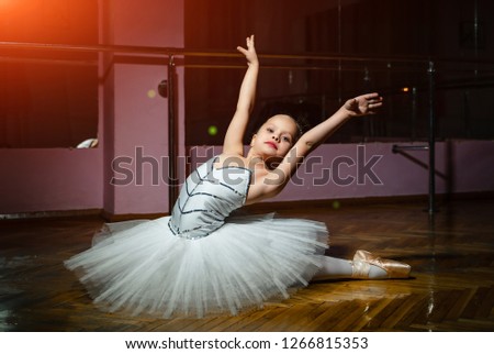 Charming little girl ballet dancer in white tutu performing ballet poses on the floor in dance studio background. Small ballerina in ballet costume showing special pose with raised arms on floor.