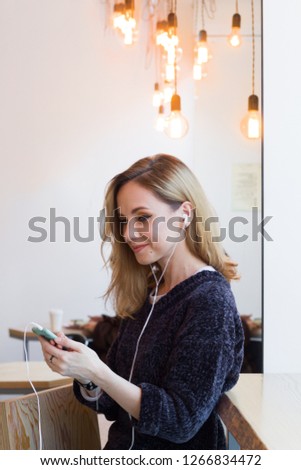 beautiful smiling young woman in gray sweater using mobile phone with earphones in cafe