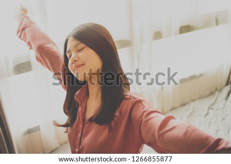 Motion image of a woman in pink pajamas stretching on a bed after a good sleep, curtains and light from the window in the background.