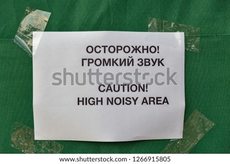 inscription in Russian and English on a white piece of paper taped by scotch on green tent background: Caution! High noisy area