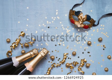 Two champagne bottles with carnival mask, confetti stars and party streamers on blue background. Flat lay of Christmas, anniversary, carnival, New Year celebration concept. Copy space, top view.