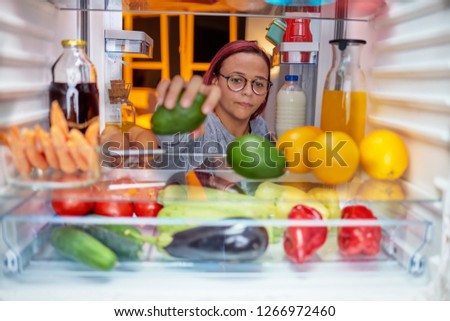 Woman standing in front of opened fridge and taking avocado. Fridge full of groceries. Picture taken from the iside of fridge.