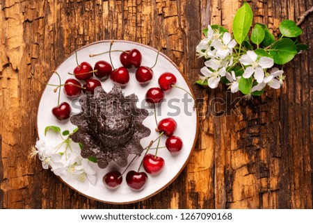 Chocolate muffin with cherry filling is served on a white plate and decorated with fresh ripe sweet cherry. Homemade baking . Background from aged textured wood. Flat layout.