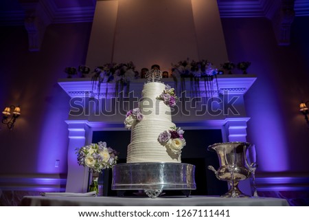 Beautiful white wedding cake with flower accents in front of mantel with purple uplights Royalty-Free Stock Photo #1267111441