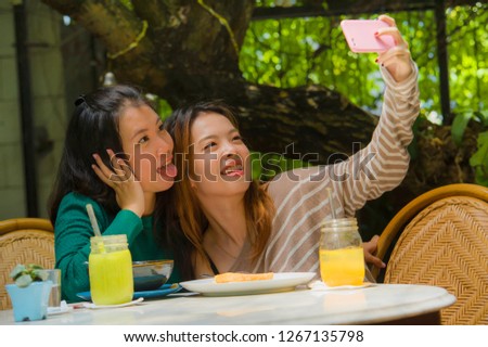 lifestyle portrait of two young happy and cute Asian Chinese girlfriends taking selfie portrait picture with mobile phone camera for using on internet social media while having healthy brunch at cafe