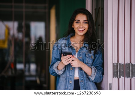 Portrait of a beautiful, tall, elegant and young Indian Asian woman texting on her smartphone. She is wearing a retro denim jacket with an 80's feel and is smiling happily.  Royalty-Free Stock Photo #1267238698