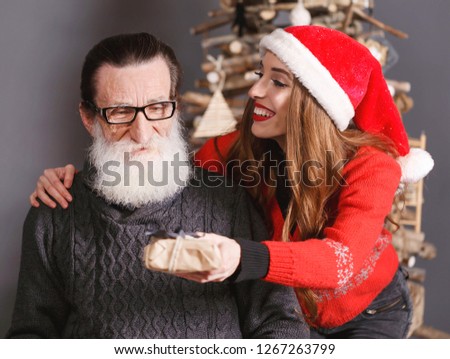 Handsome bearded senior dad wearing glasses and gray sweater accepts a gift from his young long haired daughter in the red sweater wearing Santa hat, he feeling shocked and surprised, New Year