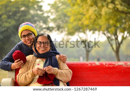 Young Indian woman with her mother knitting sweater who is sitting on a red bench in a park in New Delhi, India. Concept Mother's day
