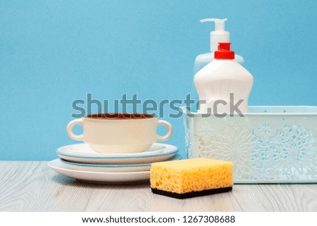 Plastic bottles of dishwashing liquid, glass and tile cleaner in basket, sponge, clean plates and bowl on blue background. Washing and cleaning concept.