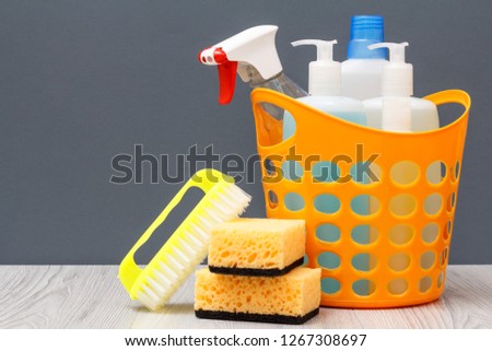 Plastic basket with bottles of dishwashing liquid, glass and tile cleaner, detergent for microwave ovens and stoves. Brushe and sponges on gray background. Washing and cleaning concept.
