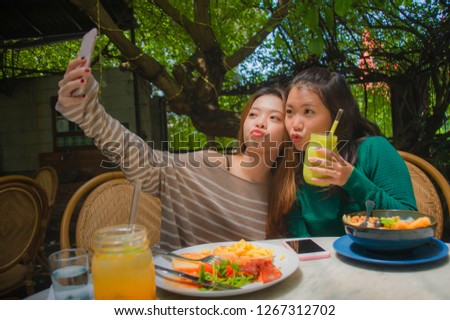 lifestyle portrait of two young happy and cute Asian Chinese girlfriends taking selfie portrait picture with mobile phone camera for using on internet social media while having healthy brunch at cafe