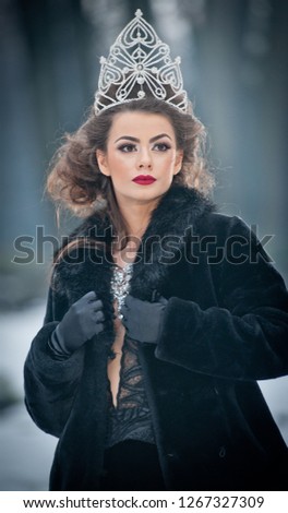 Beautiful fairy-tale winter queen in the forest with sparkling tiara and Elegant black fur coat. Fluffy snow flakes.