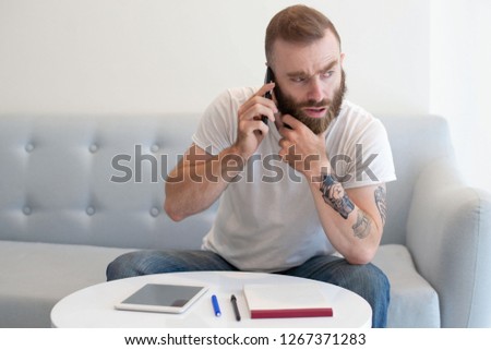 Frowning bearded freelancer talking on phone in lounge or waiting room. Young man in white t-shirt and jeans sitting on couch and speaking on cell. Business anywhere concept