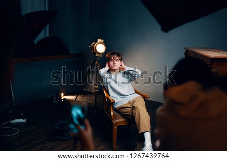 The guy in a sweater and pants sits on a wooden chair and poses for a photographer in a room with a searchlight            