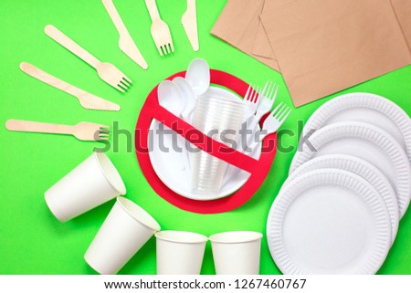 No use symbol in red forbidden sign with plastic dishes around disposable paper dishes and a wooden fork and knife on green background. Environmental concept. Ban single use plastic.