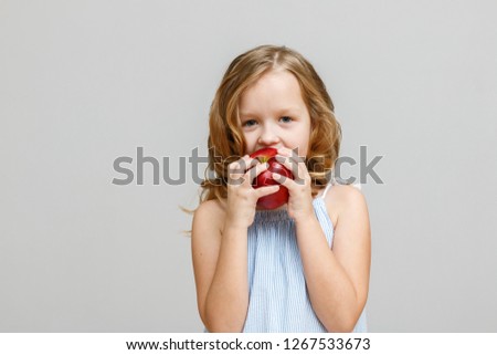 Portrait of a happy smiling little blonde girl on a gray background. Baby eating red apple