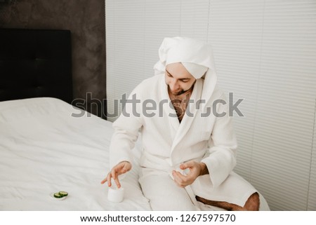 Relaxed bearded young man in a towel on his head and white coat with moisturizer on the face. Photo of smiling man on beige background. Grooming himself after shower
