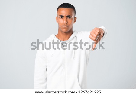 Dark-skinned young man with white sweatshirt showing thumb down sign with negative expression on grey background