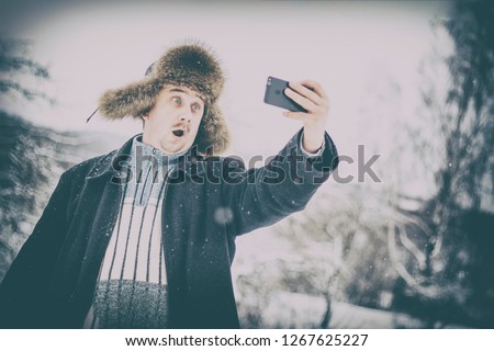A young man is surprised at the camera, makes a selfie in a snowy winter forest. the guy on the street. on it hats made of natural fur. there is toning.