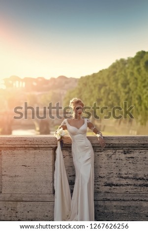Beautiful young blonde bride with elegant hairdo in white wedding dress with long train holding bouquet of calla flowers with blue ribbon standing on bridge on landscape background, vertical picture