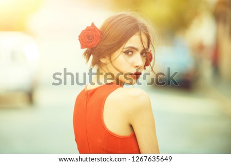 woman with red lips, makeup on cute young face and roses in brunette hair posing outdoors on sunny, summer day on blurred background. Visage. Beauty and hairdressing salon