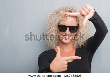 Young blonde woman with curly hair over grunge grey background smiling making frame with hands and fingers with happy face. Creativity and photography concept.