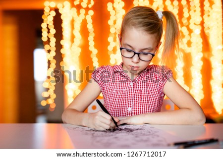 Blonde girl in the pink dress and big black glasses drawing santa claus. Christmas and New Year theme, yellow bokeh.
