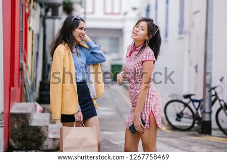 Two young and attractive Asian women talk animatedly with one another in an alley during the day. They are both beautiful and wearing retro 80's clothes. One is Indian, the other Chinese. 