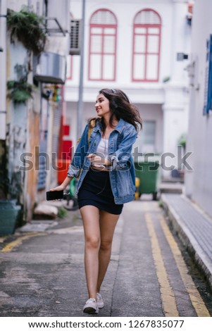 Portrait of a young and attractive Indian Asian woman in a denim jacket and yellow backpack enjoying music on her headphones. She is smiling as dancing as she walks down an alley on her way somewhere.