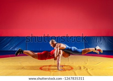 Two strong wrestlers in blue and red wrestling tights are wrestlng and do push-ups and gymnastic exercises in pairs, plank in the gym. Young man doing grapple.