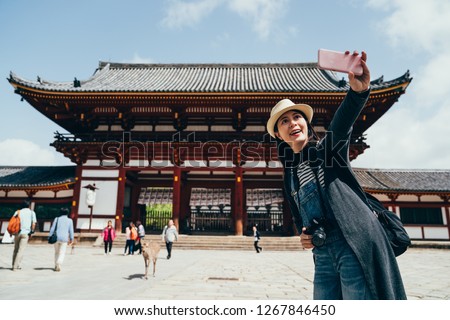 Lady tourist in straw hat visiting todaiji nara Japan taking selfie by smart phone. young girl travel backpacker using cellphone making self portrait with traditional japanese temple in background.