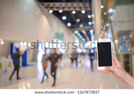 Hand holding mobile phone with abstract blurred background image and bokeh light of crowd people at cars exhibition show, internet and social network concept