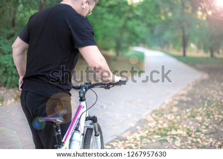 Young caucasian man holds white bicycle on the path of abandoned park, black casual wear. Dark hair, earring and earphones, round golden glasses. Outdoors, copy space, lenz flair effect,early autumn. 