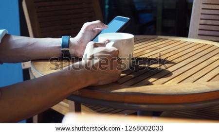 Closeup of Asian male hands playing smartphone and drinking coffee at an outdoor table with morning sunlight.(Selected focus)