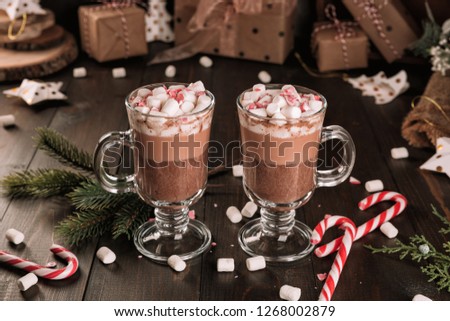 Cup of hot chocolate cocoa drink with a marshmallows and candy cane on wooden table. Two glasses of a sweet winter beverage and fir-tree branch. 