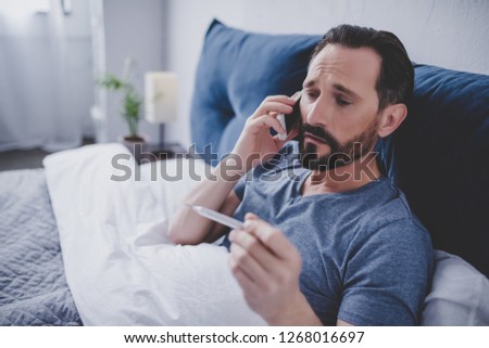 portrait of sick man holding thermometer and calling a doctor while sitting on the bed Royalty-Free Stock Photo #1268016697