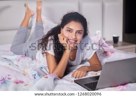 Picture of a beautiful woman smiling at the camera while lying with a laptop on the bed. Shot at home