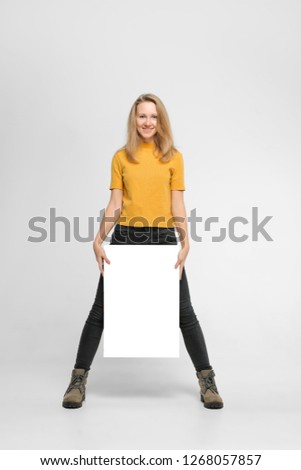 Positive laughing woman wearing black jeans and yellow t-shirt with blondie hair, toothy smile is holding white big mockup poster near her legs isolated on white background
