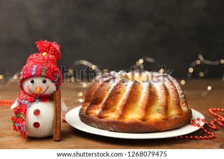 Christmas cake on white plate and toy smiling snowman in red hat and scarf on wooden table on black background with yellow lights.